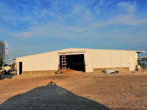 Large cream-colored industrial warehouse building with open bay door under blue sky on red dirt lot