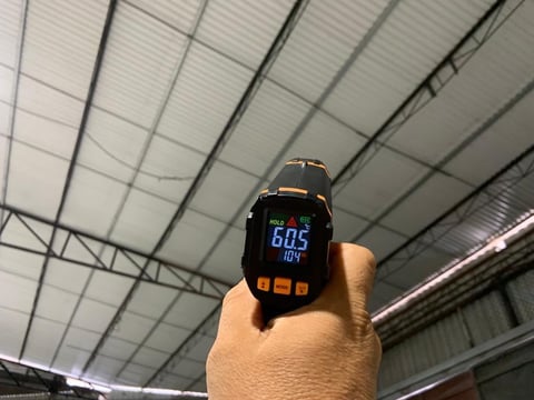 Hand holding infrared thermometer displaying 60.5 degrees inside an industrial building with metal ceiling