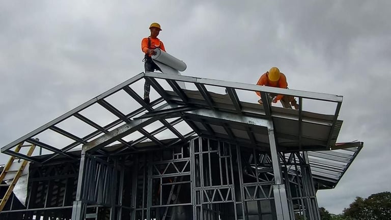 Two construction workers installing roofing materials on a metal frame structure against a cloudy sky