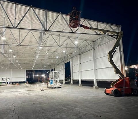 Large illuminated industrial warehouse with metal framework, open bay doors, and red aerial lift equipment parked inside at night